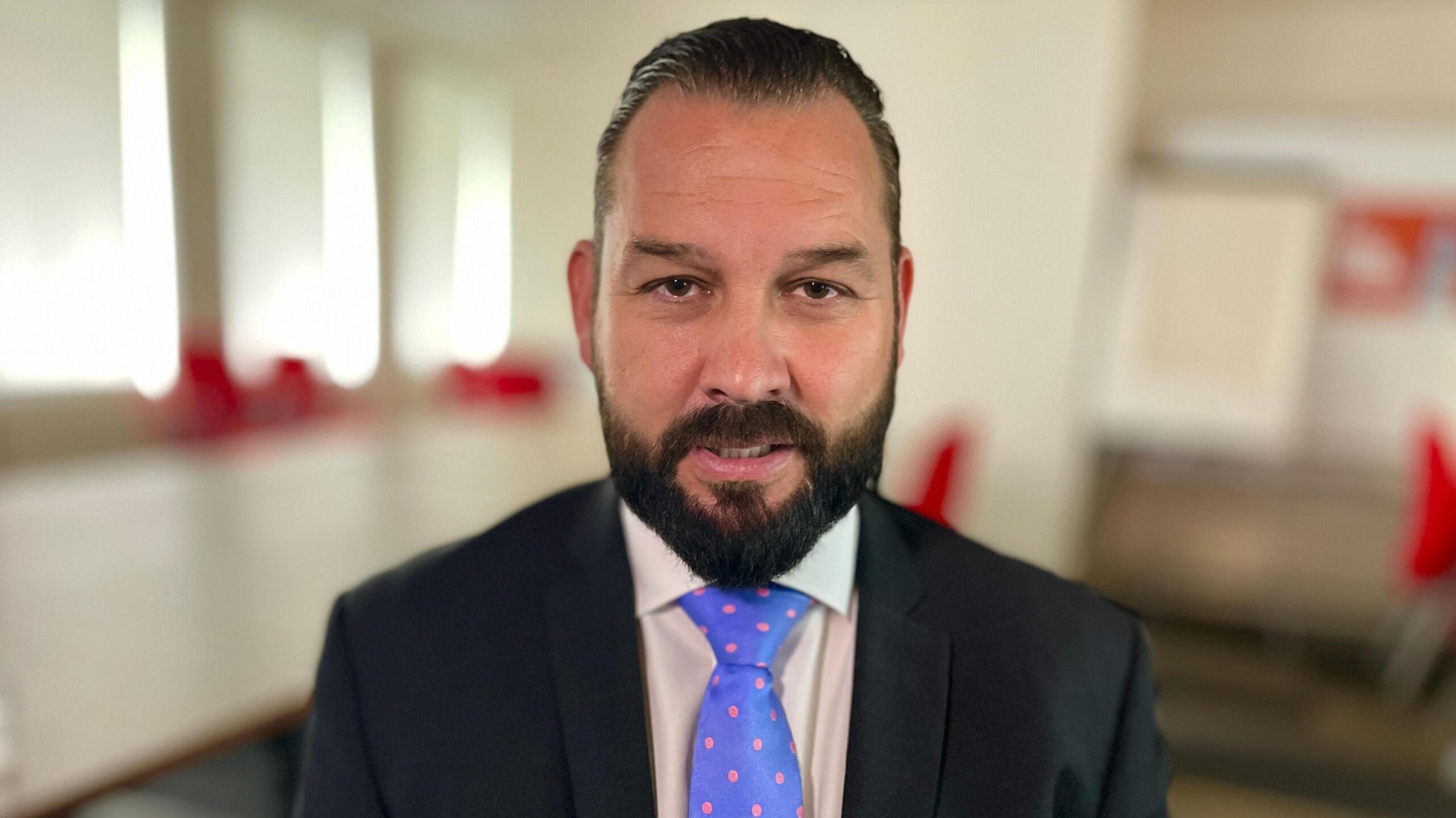 A man smiles at the camera as he sits inside a room. The background behind him is blurred. He has dark hair that has been slicked back and has dark facial hair. He is wearing a black suit jacket with a white shirt understand, and a blue tie with pink spots on it.