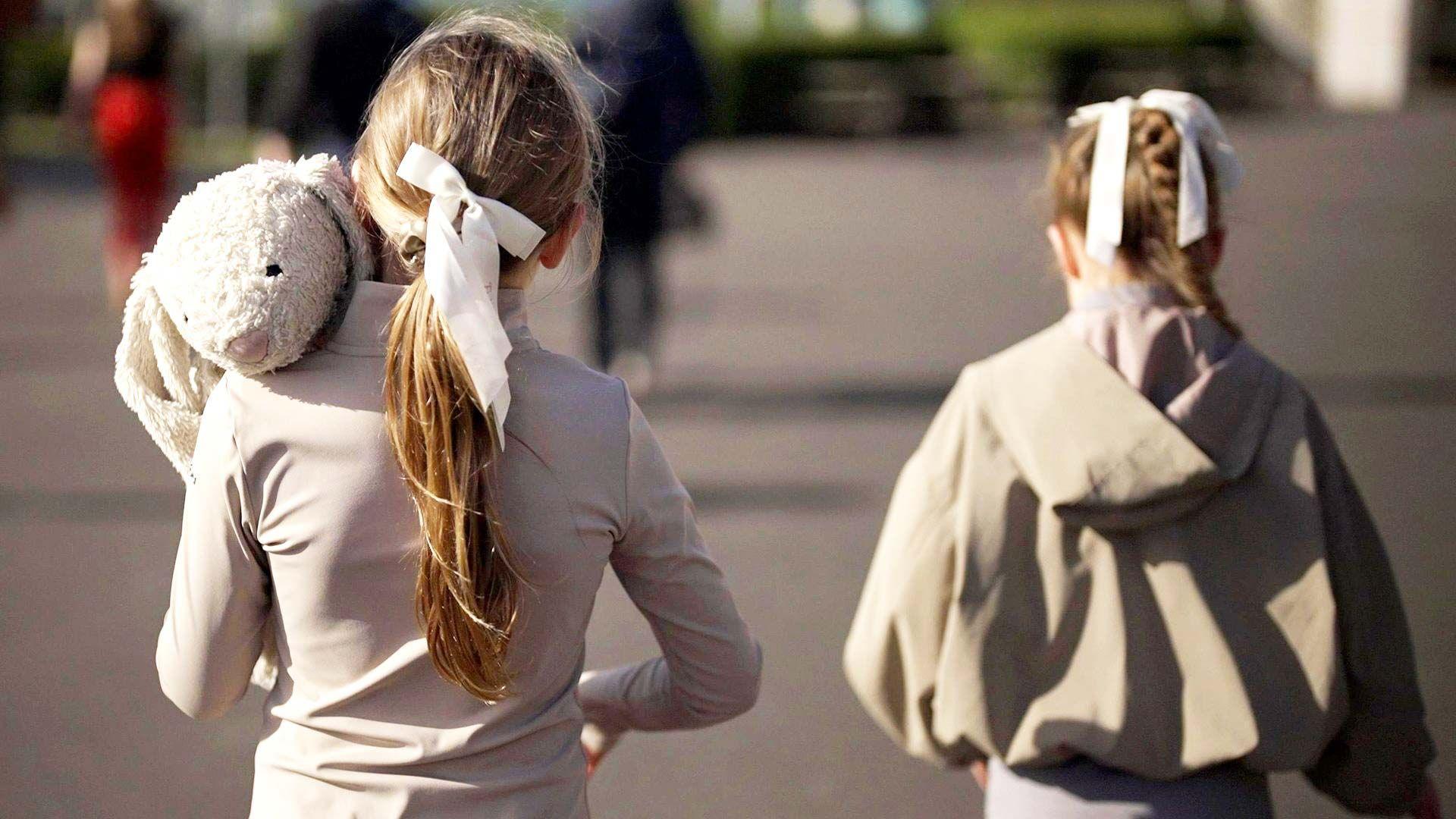 Two girls seen from behind wearing beige and cream clothing  with white ribbons in their hair. One is holding  a white rabbit soft toy