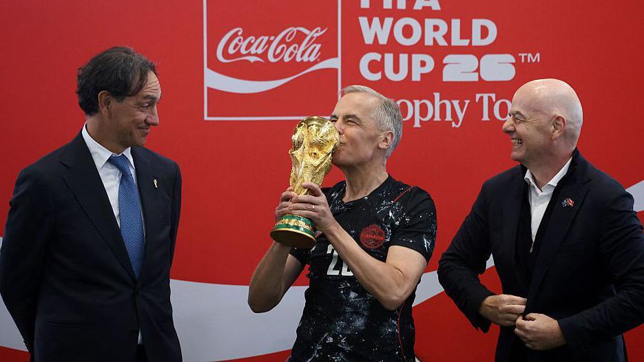 Canada's Prime Minister Mark Carney, in a black Canada jersey, kisses the FIFA World Cup trophy, as FIFA President Giovanni Infantino and Italian football coach Alessandro Nesta, both in suits, look on, amused.