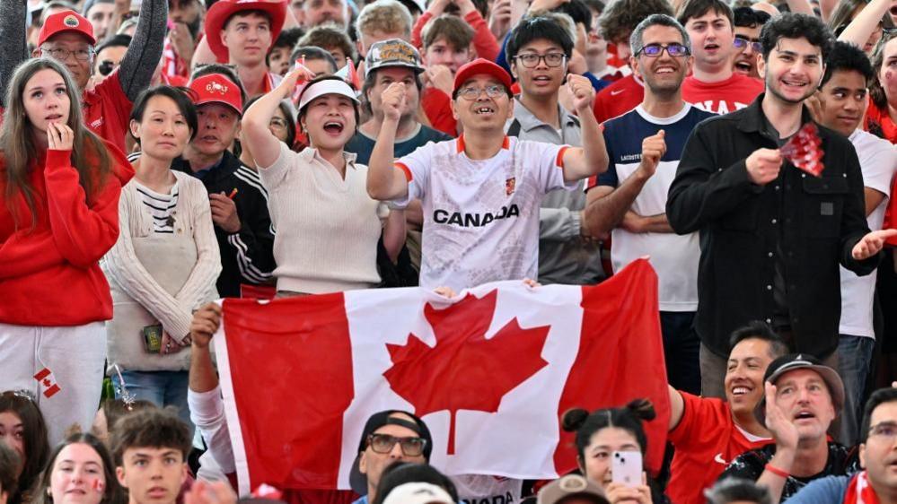 A crowd of Canada fans, many in red and white, react to a play by Canada. Two are holding up a Canadian flag.