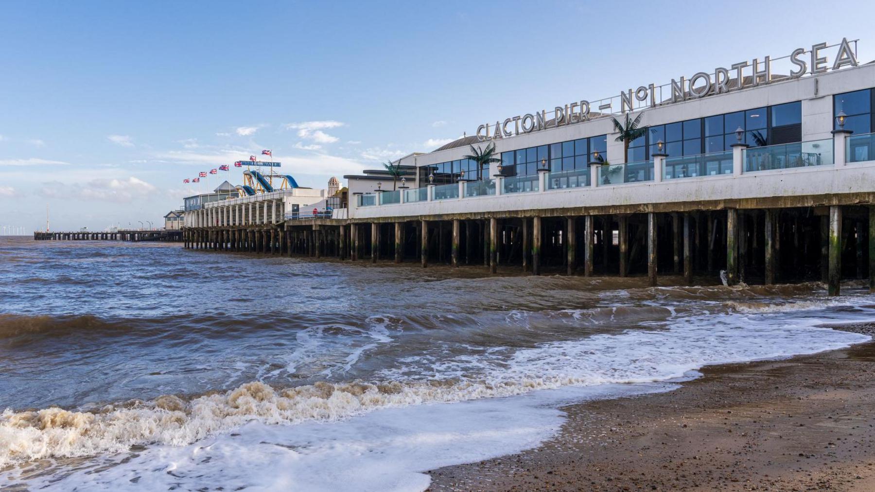 A side-angle view of Clacton Pier extending over choppy, brown ocean waves onto a sandy beach under a partly cloudy sky. The large building on the pier features large windows and a sign on top that reads 'CLACTON PIER - NO. 1 NORTH SEA,' with amusement park rides visible further down the deck