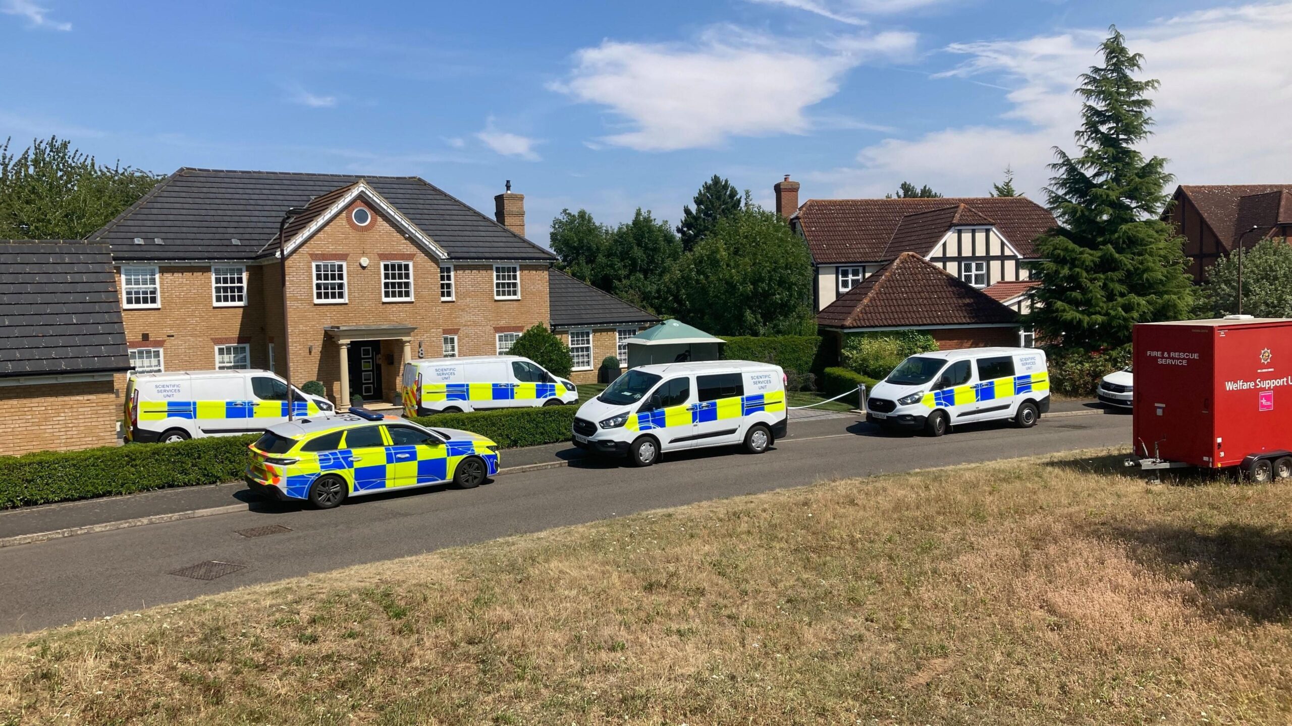 About six police vehicles parked outside a large modern, two-storey brick house. Next door on the right is a mock Tudor house - both have separate garages in front of them. There is also a red vehicle/trailer with "Fire & Rescue Service" and  "Welfare Support..." written on it.