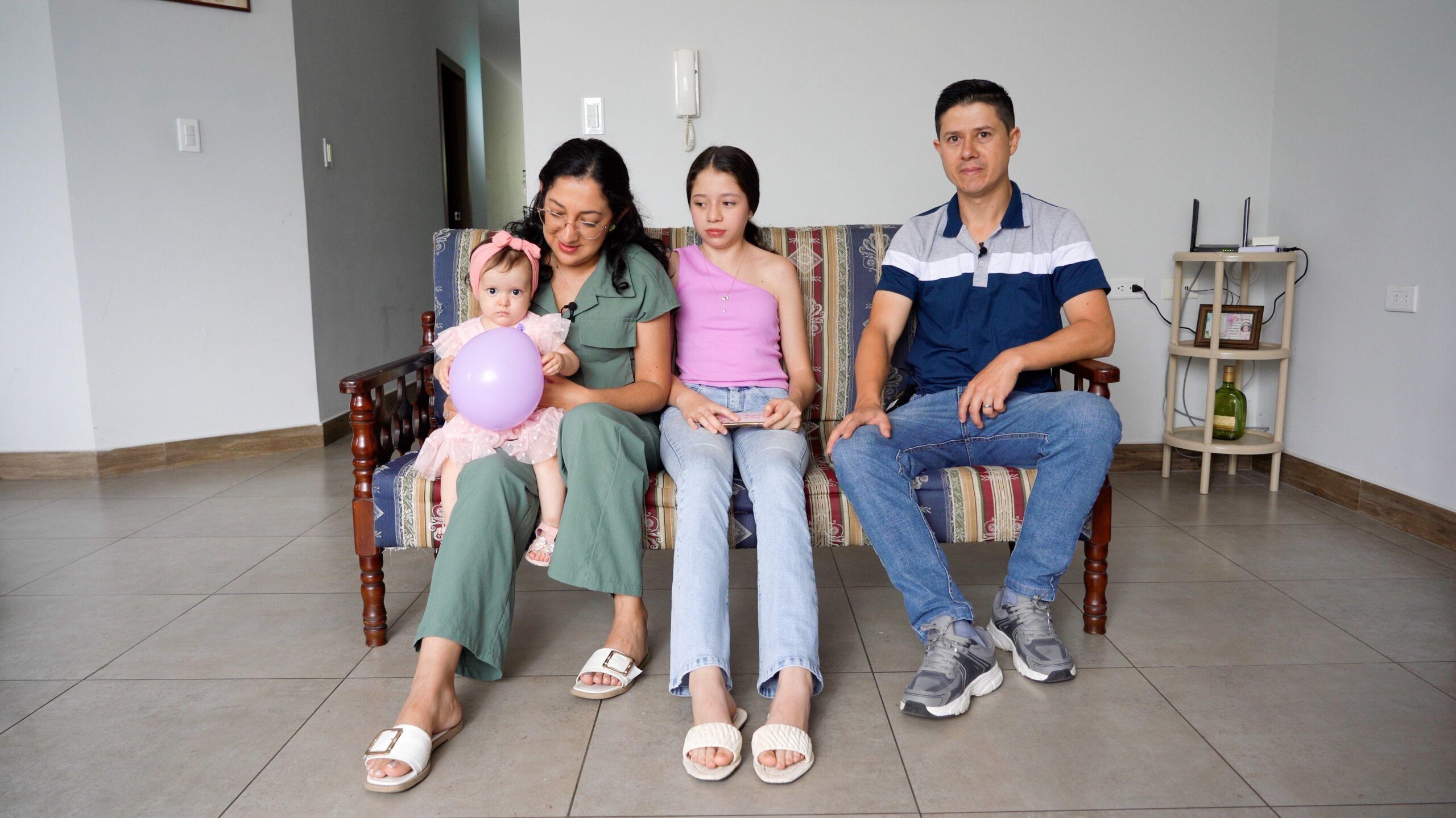 Camila pictured with her family who are sitting on a sofa in a living room with a tiled floor. She is wearing a pink dress and hair band and is holding a purple balloon. She is sitting on the knee of her mum who has long dark hair and glasses, and is wearing a green jumpsuit. Her sister is in the middle with a purple off-the-shoulder top and jeans, next to her dad who has short dark hair, a blue polo t-shirt and jeans.