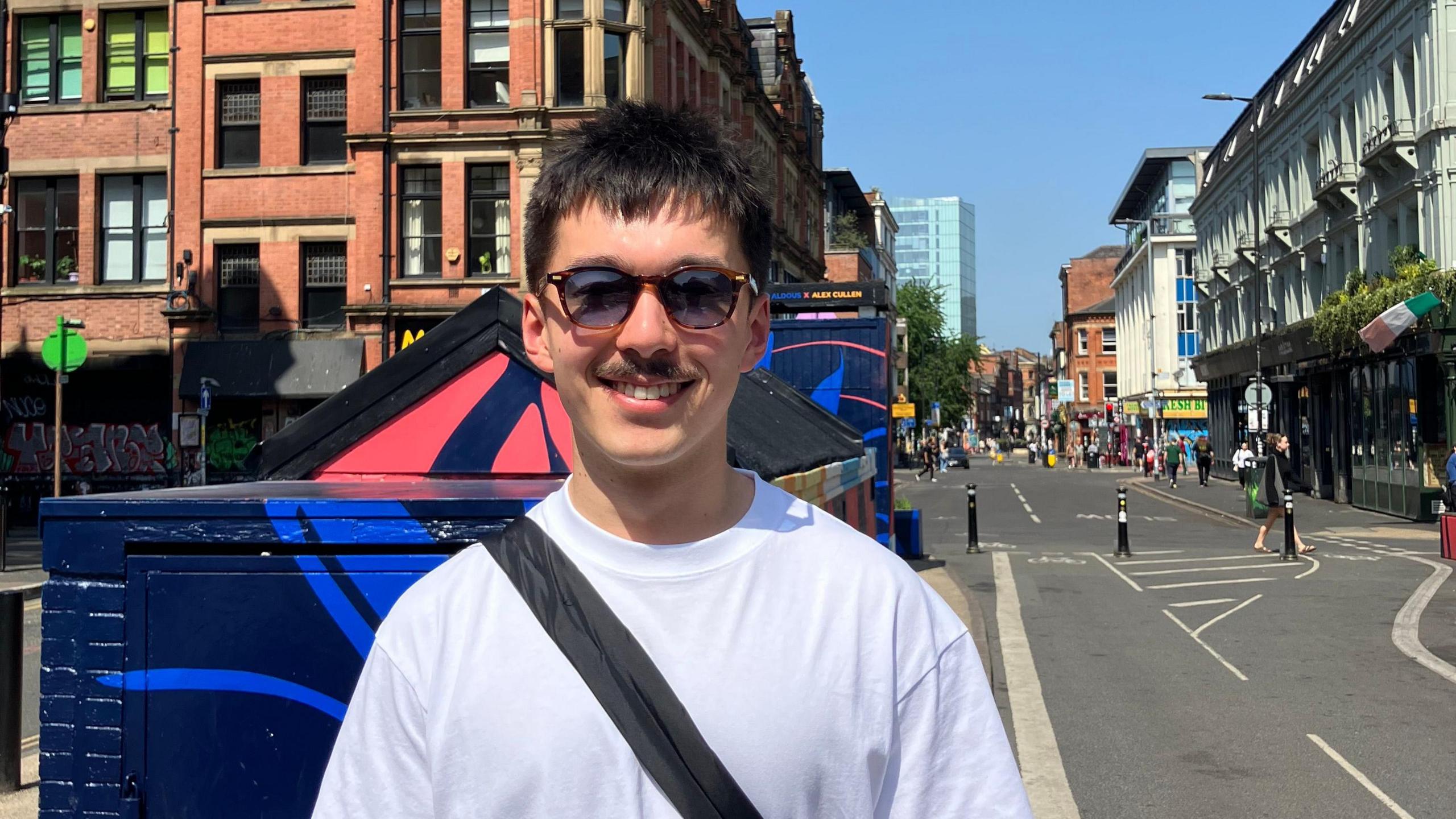 Harry in the foreground wearing a white T-shirt and sunglasses, with a black crossbody bag, standing on a pavement beside a blue-painted utility structure. Behind, a two-lane street has white road markings and a separated section with short black bollards. Pedestrians walk along pavements on both sides. Red-brick and light-coloured buildings line the street, with shopfronts and signage visible. A lamppost stands above the pavement, and taller modern buildings appear in the distance under a clear blue sky.