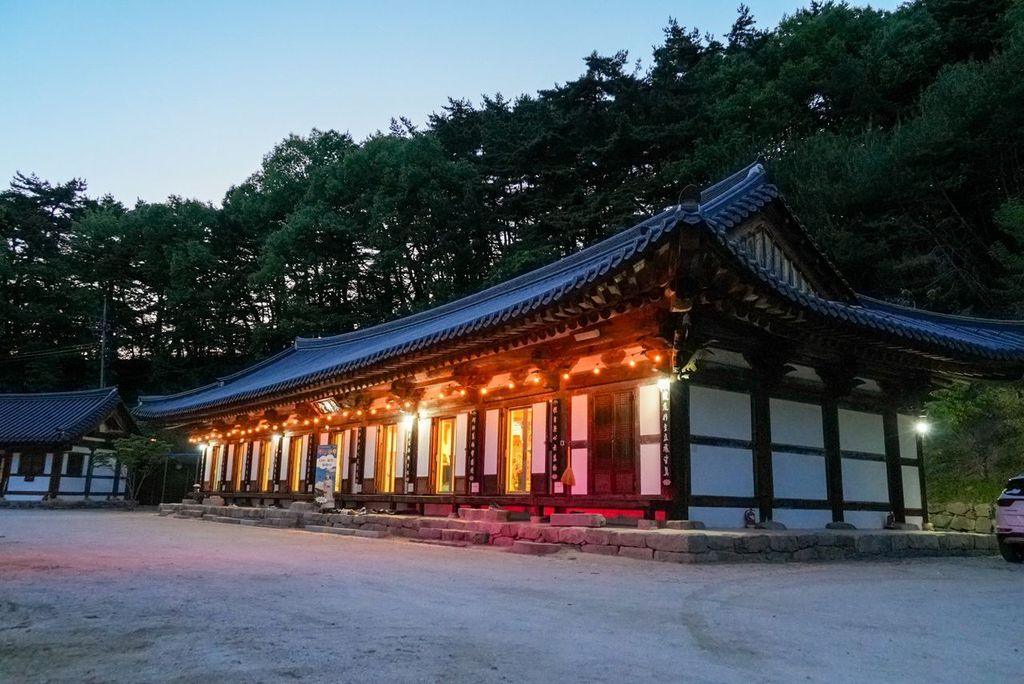Traditional Korean temple illuminated with warm lights at dusk, set in a quiet courtyard at the edge of a forested hillside