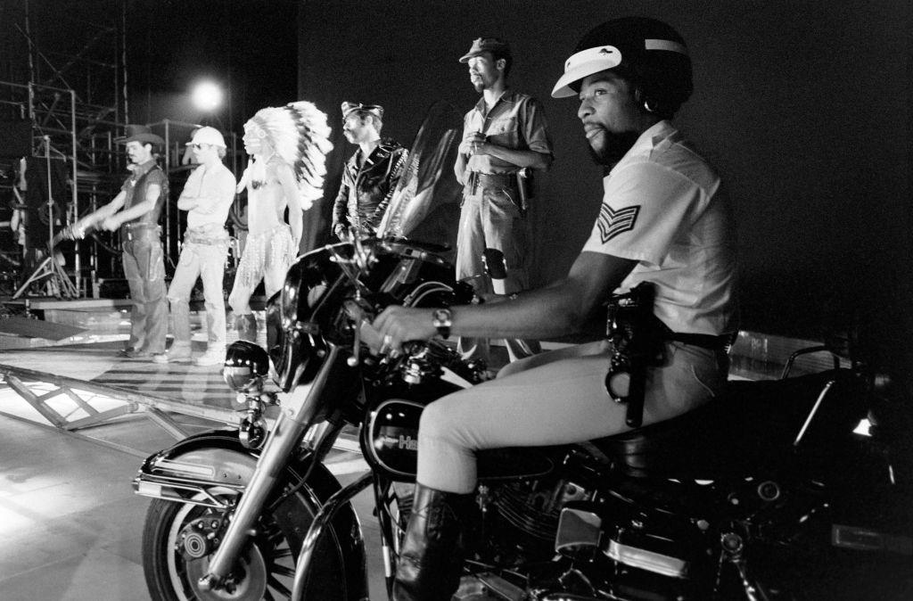Victor Willis sits on a motorcycle as the rest of the Village People perform on stage