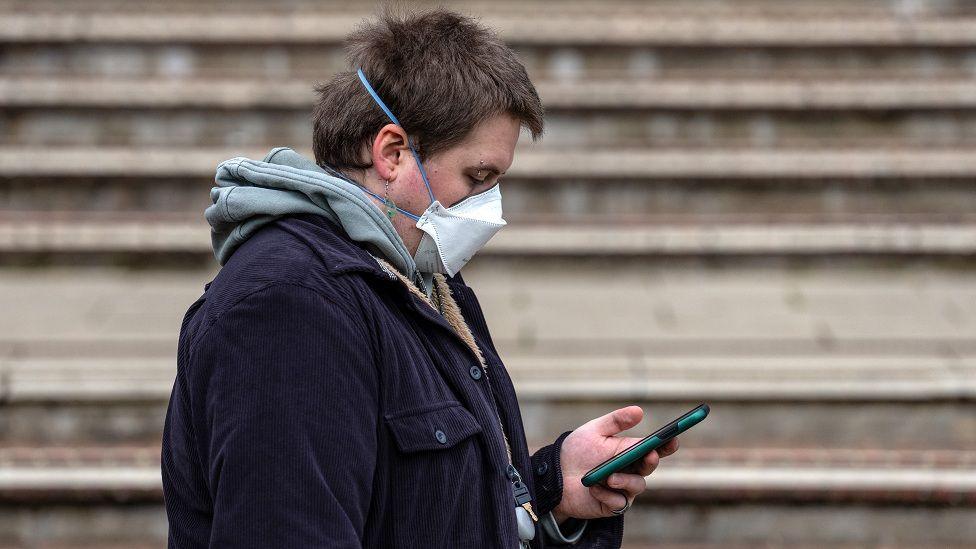 A young person wearing a hoodie, jacket and a mask across his face reads his phone.