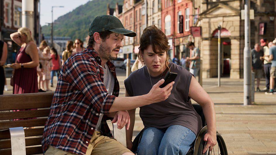 Iwan Rheon and Ruth Madeley filming on set in a town centre, both looking at the same mobile phone screen in summery, sunshine conditions