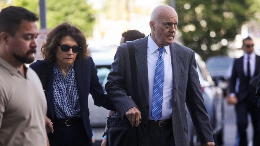 A balding man in a grey suit holds the hand of a woman in a navy blue suit who is wearing sunglasses as both are walking