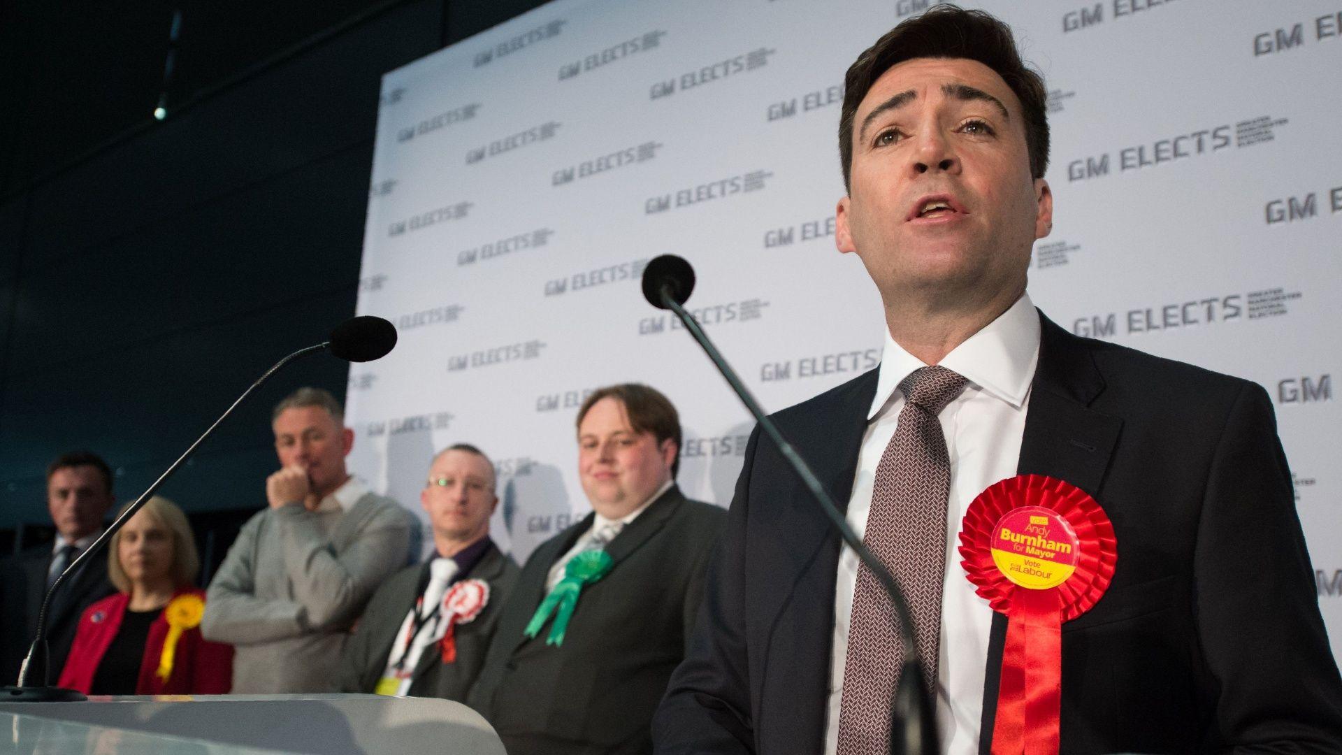 Andy Burnham, wearing a black suit, white shirt and burgundy tie with a red rosette on his lapel, speaks into a microphone next to a number of smartly-dressed fellow candidates for Greater Manchester mayor, in front of a wall with signs on it reading 'GM Elects'
