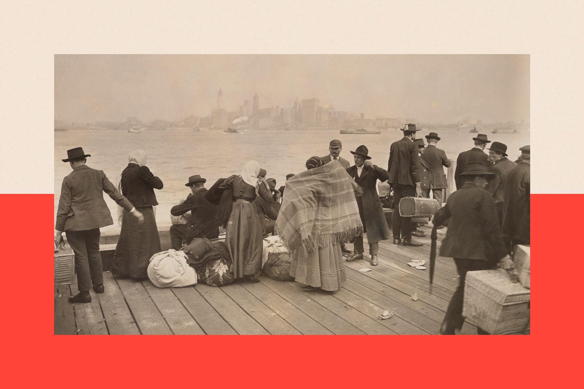 A group of immigrants wait at Ellis Island to be taken to Manhattan. They wear cloaks, hats and coats.