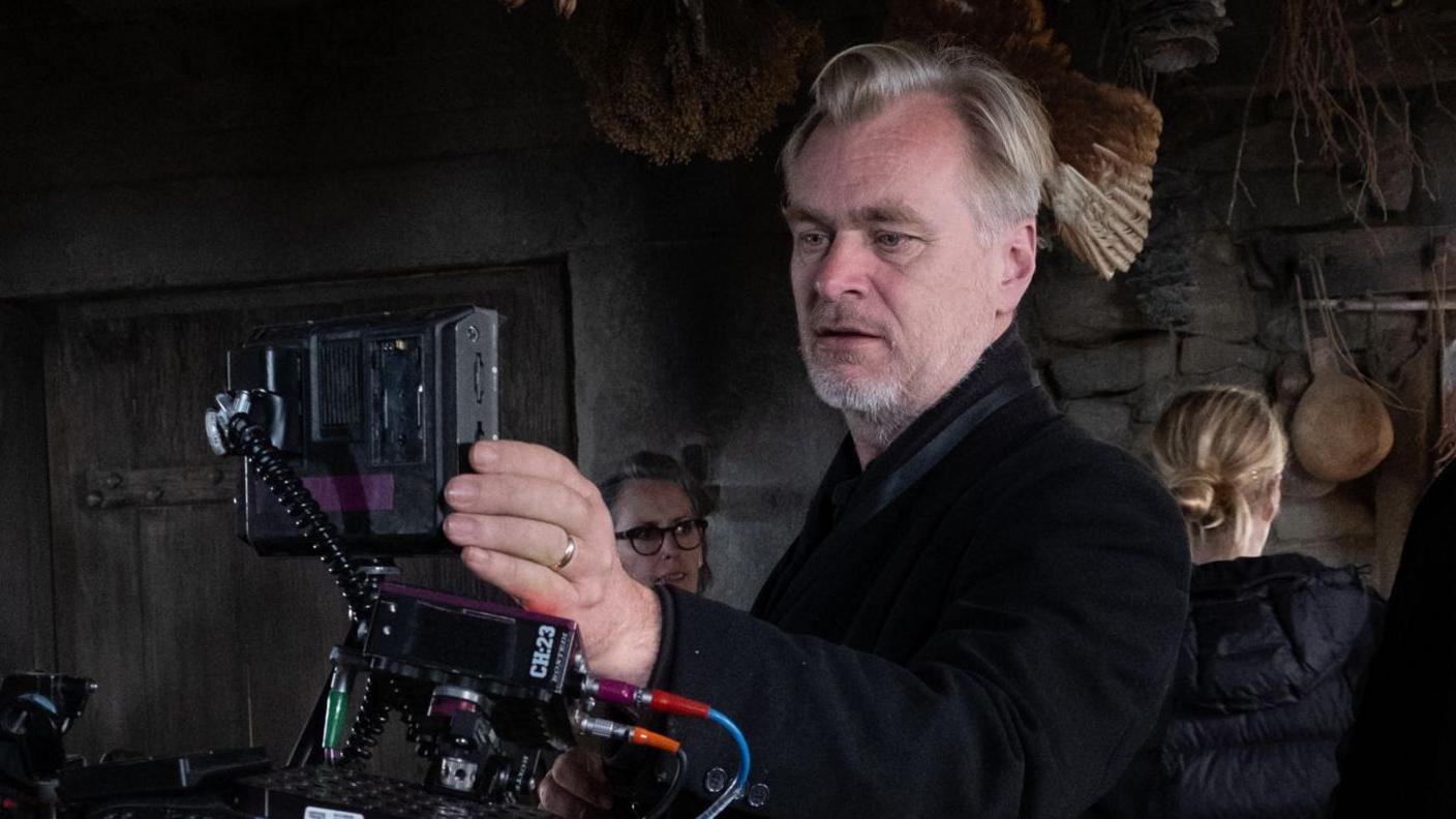 Christopher Nolan in the centre of the photo and is looking through an Imax camera. He is wearing a black jacket.