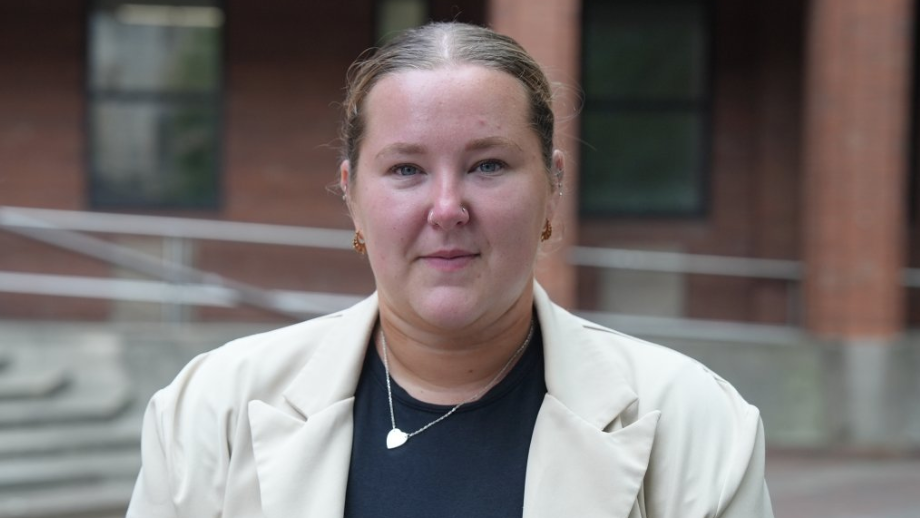 A woman with tied-back brown hair stands outside a court building. She is wearing a cream jacket over a navy top. She is donning a hear-shaped locket.