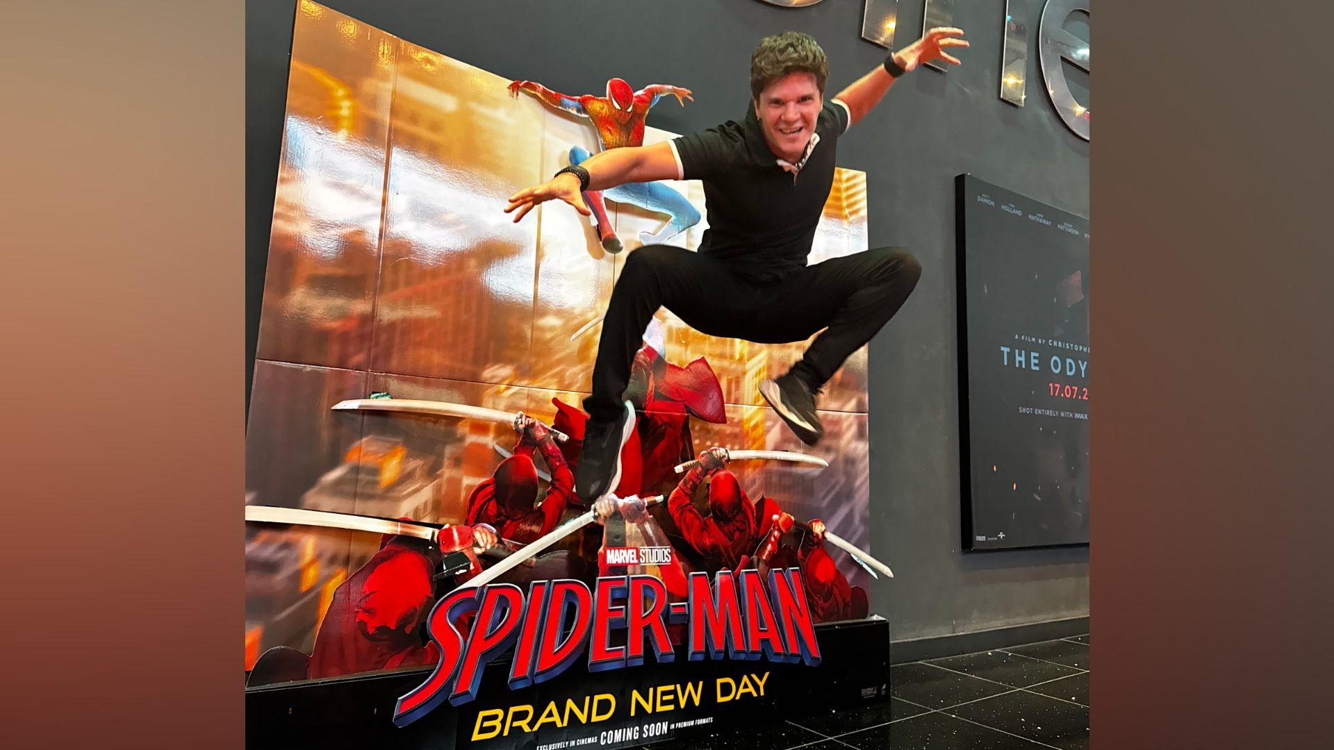 A man jumps up and strikes a pose with his arms outstretched. He is in front of a sign reading 'Spider-Man - Brand New Day' against a grey cinema lobby wall