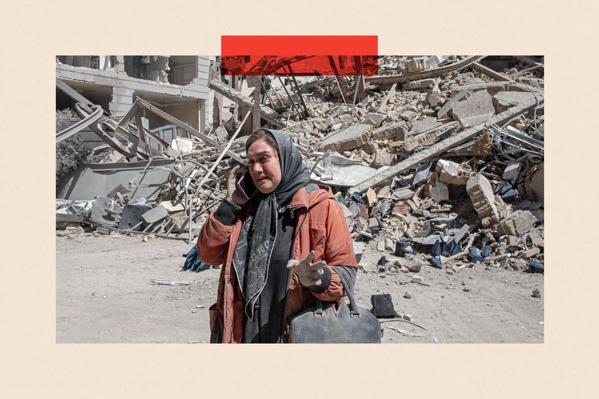 A woman in an orange jacket and grey headscarf talks on the phone in front of a destroyed building