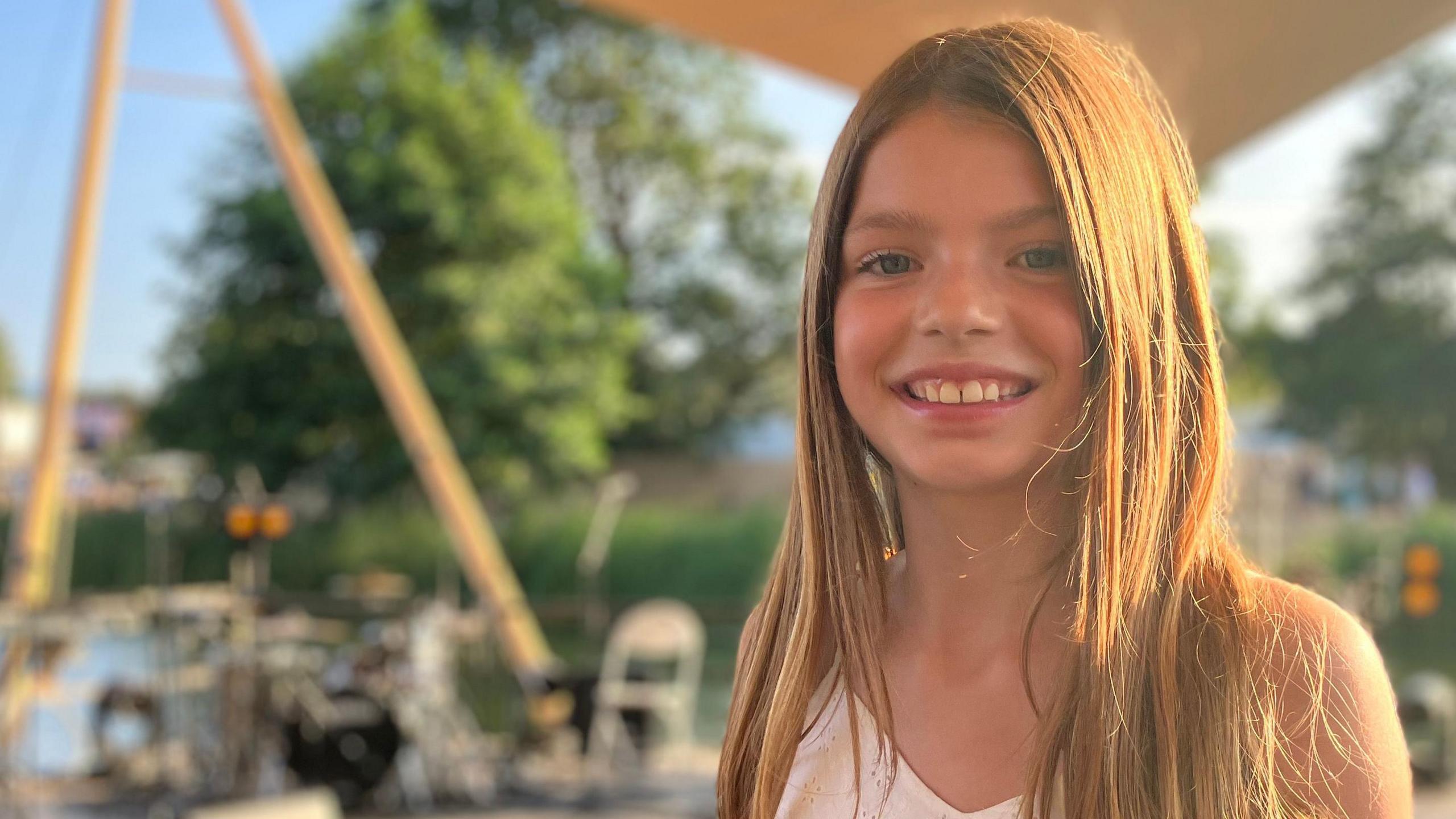 A 10 year old girl with long hair stands on an outdoor stage.