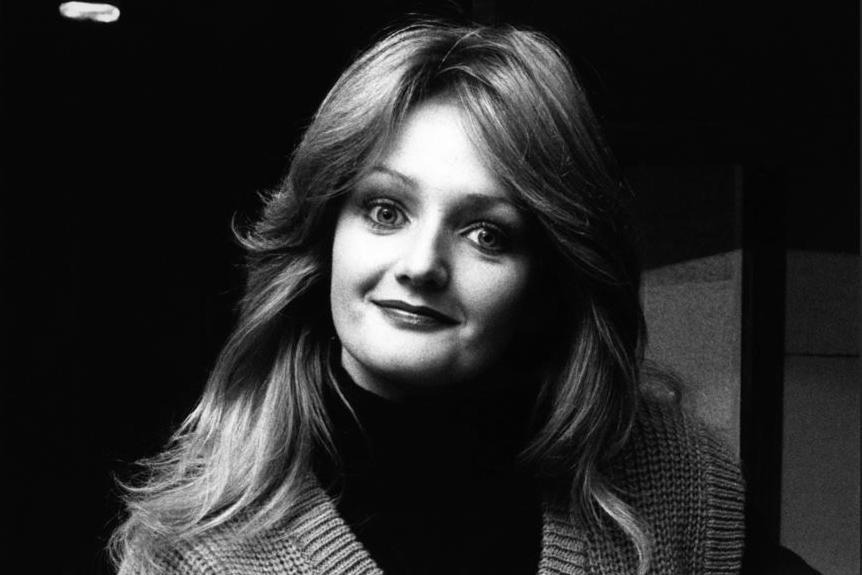 Black and white image of a young Bonnie Tyler, with long hair flowing past her shoulders. She wears a v-check woollen sweater vest with a long sleeve t-shirt underneath.