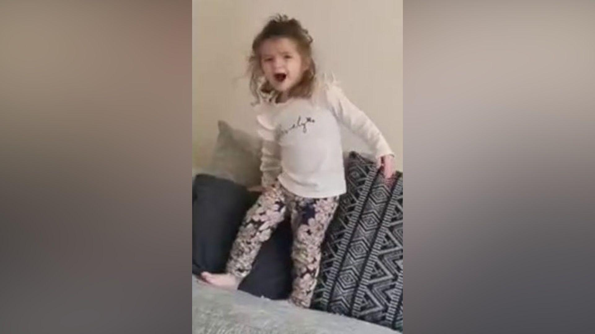 Still from a video showing Isabelle running around on a grey sofa. She is smiling and excited. She is wearing flowery leggings and a white shirt and has shoulder length brown haair.