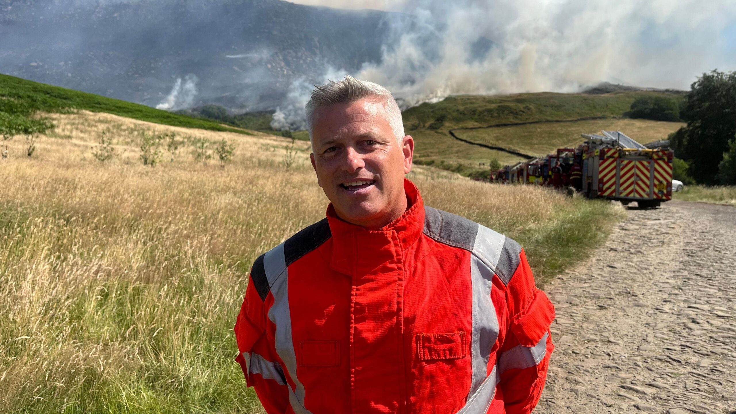 Paul Fearnhead has short grey hair and is wearing a fluorescent fire service uniform is standing on a dirt track while being interviewed. Smoke can be seen behind him.