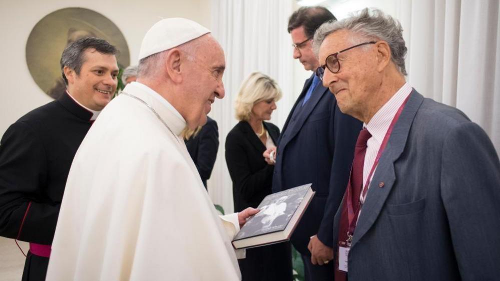 Pope Francis, dressed all in white, is holding the book and smiling as he talks to David Willey, who is also smiling.