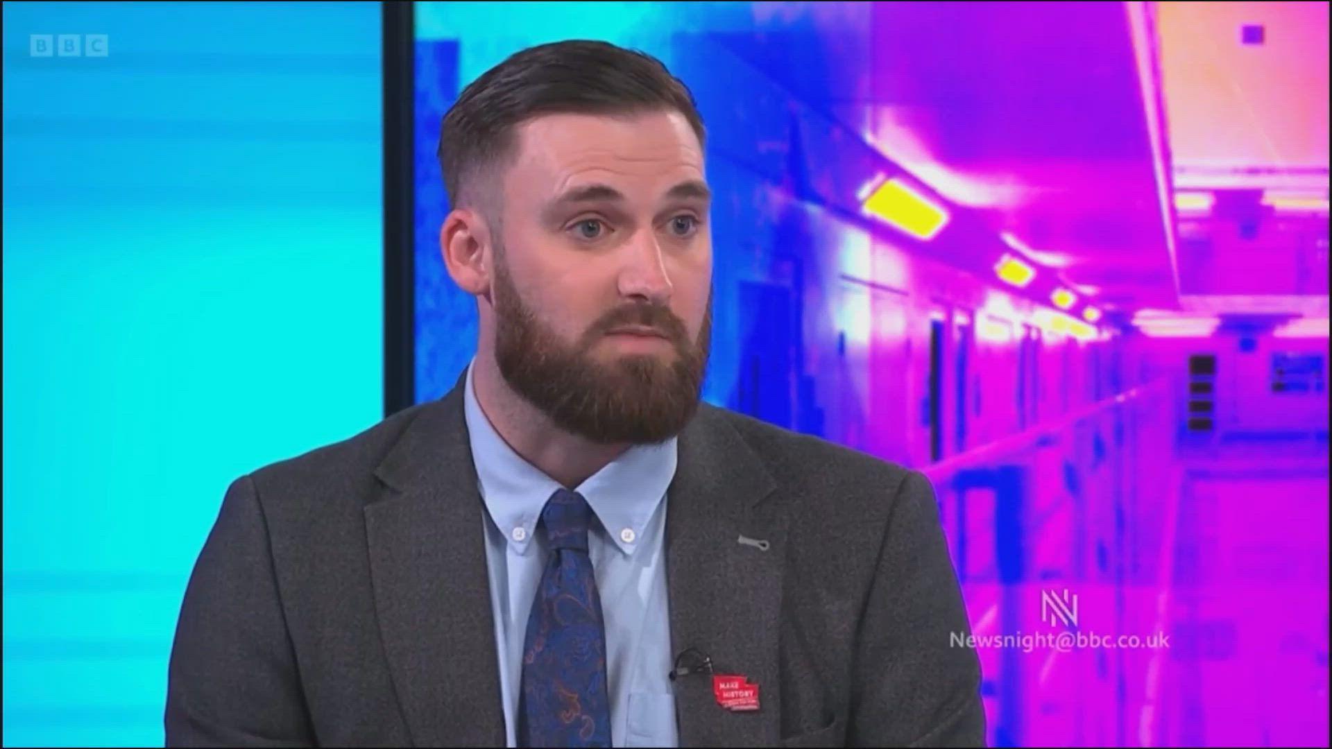 A man in his 30s, with dark brown hair and a dark brown beard, sits in a television studio wearing a dark grey suit, light blue shirt and dark blue tie.