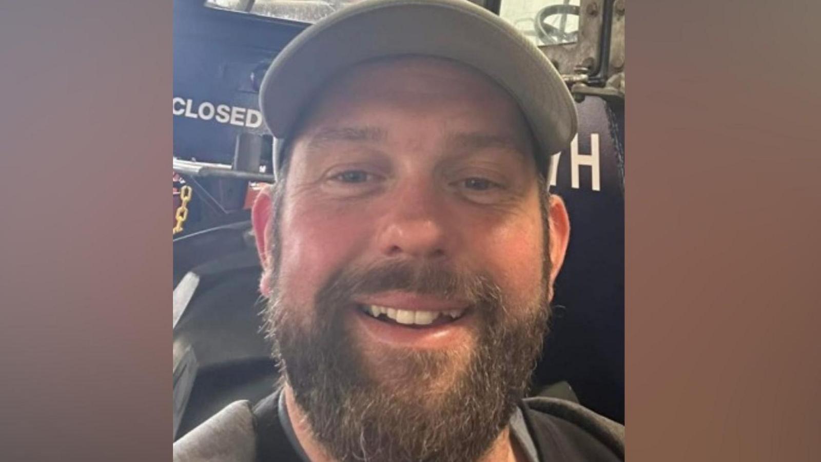 James Desborough is smiling. He has a brown beard and is standing in front of a blue tractor. He has a grey baseball cap on.