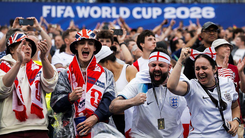 Fans inside a football stadium draped in red and white scarves and wearing white football shirts.
