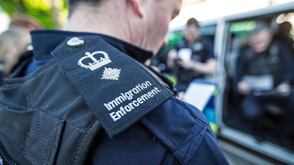 A close up of the lapel badge of an immigration enforcement officer. Other officers are blurred in the background.