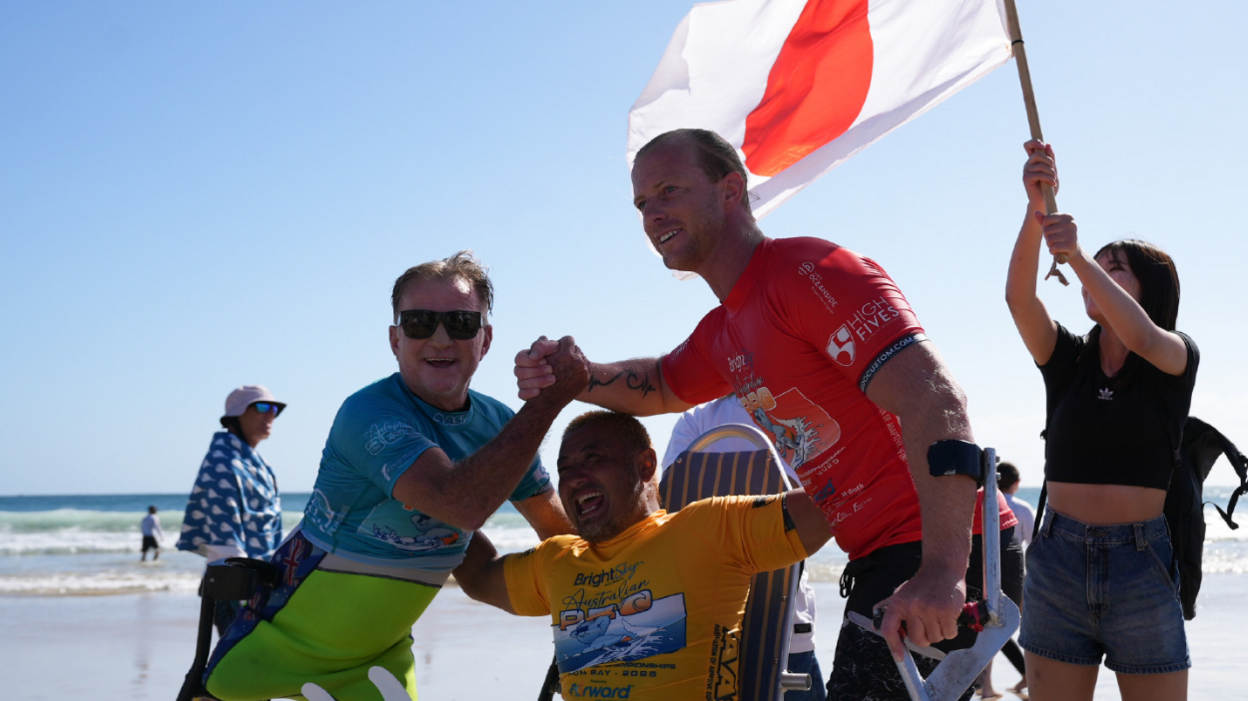 In celebratory mood: Llywelyn Williams and two other men, all wearing wetsuits and surf gear, huddle up for what looks like a group photo with the blue sea and white waves at their backs