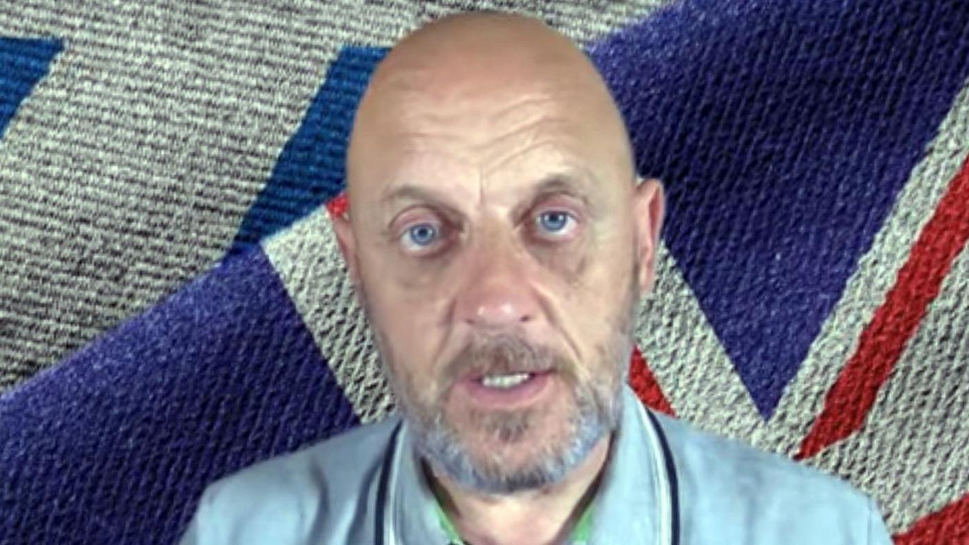 A bald and bearded man with blue eyes sitting in front of a Union flag