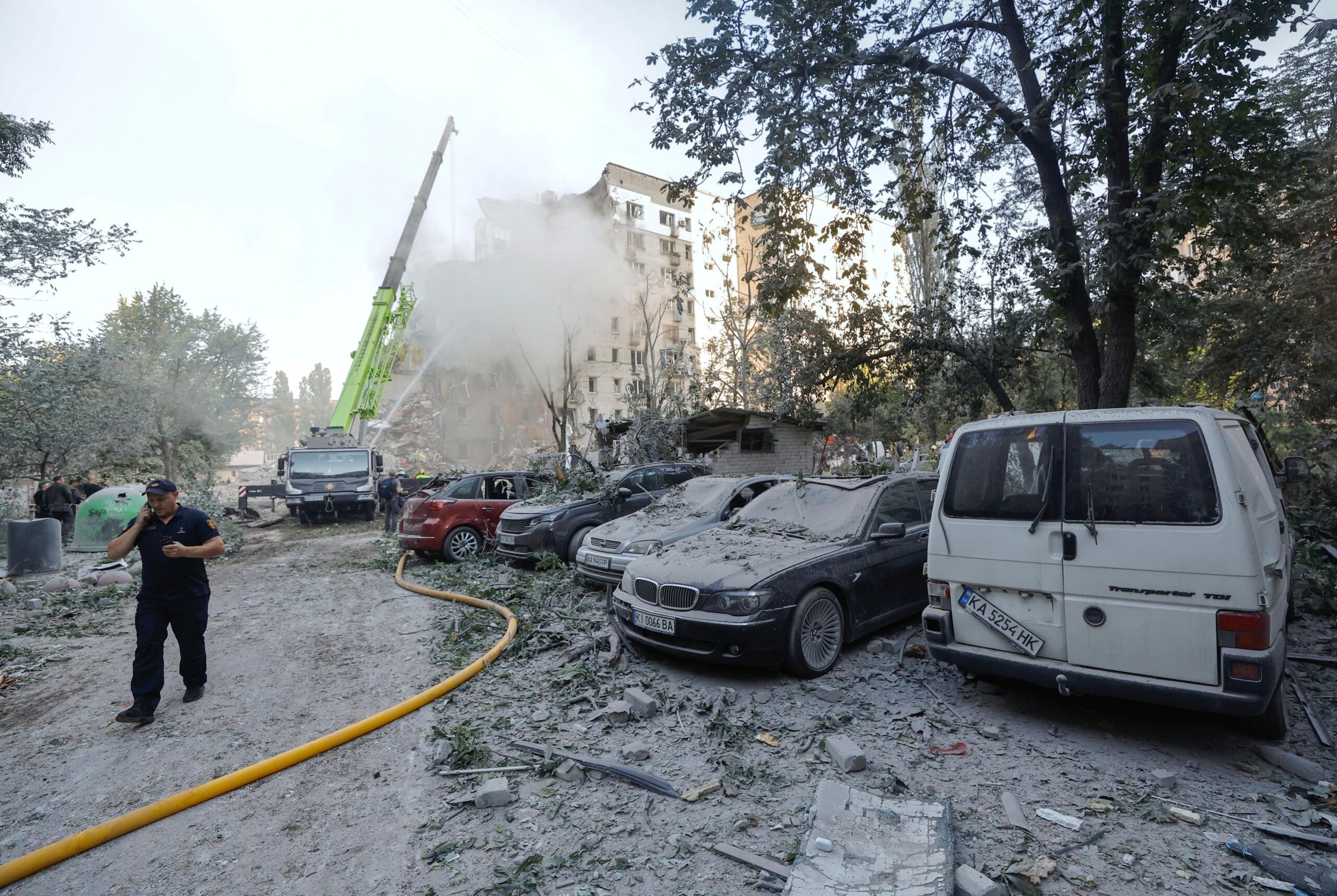 A rescue worker wearing a navy blue uniform and cap is talking on the phone while walking past a row of five cars covered in dust and debris. In the distanct there is a crane and a hose shooting water pointing towards a building covered by a cloud of smoke and dust that has been partially damaged by a missile strike.