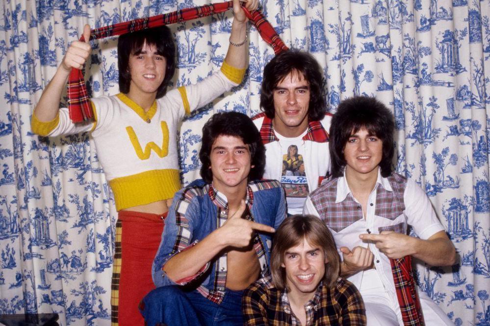 The Bay City Rollers pictured in the 1970s. They are five young men with spikey and shoulder-length hair, wearing shirts incorporating tartan. One of them is holding up a tartan scarf. They are standing against a blue and white Willow pattern curtain