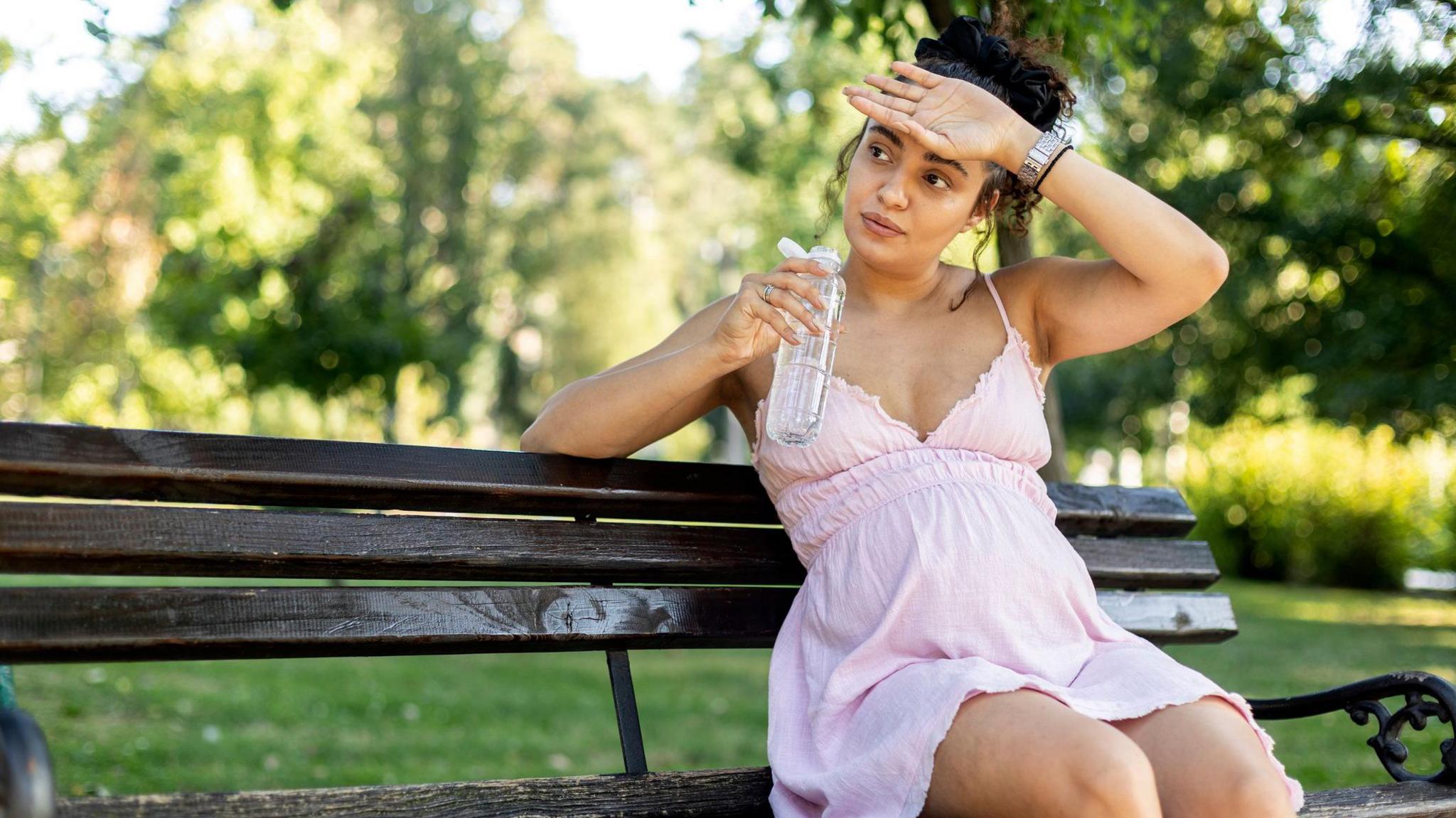 Pregnant woman sitting on a park bench in a light pink dress, holding a bottle of water.