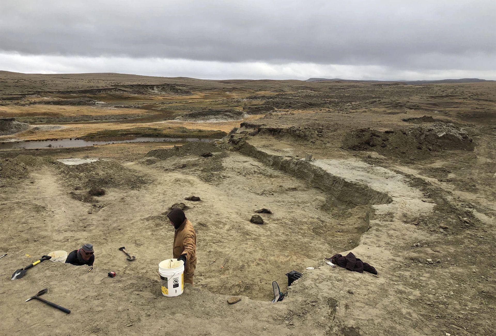 A wide view shows the barren brown landscape of the Badlands in South Dakota, with two people in the foreground on a dig with tools around them, on a cloudy grey day.