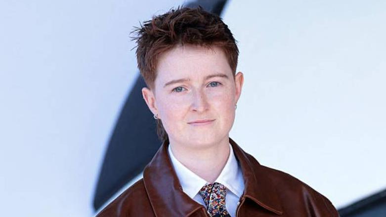 A person with short reddish hair wearing a white shirt, colourful tie and a red leather jacket