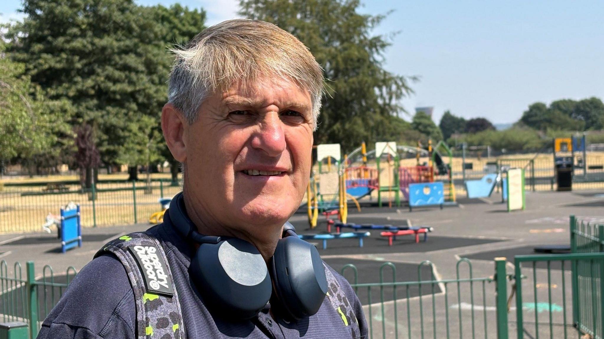 A man in his 60s with greying brown hair has large earphones around his neck and a backpack on. He is wearing a purple t shirt.