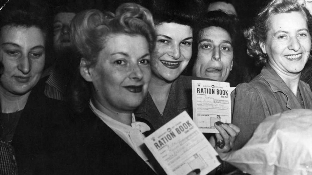This black-and-white photograph shows a group of people standing closely together in what appears to be an indoor public setting. Several individuals in the foreground are holding up documents clearly labelled “RATION BOOK”. The crowd fills most of the frame, and the image focuses on the ration books as key objects, suggesting a scene connected to food rationing or distribution.