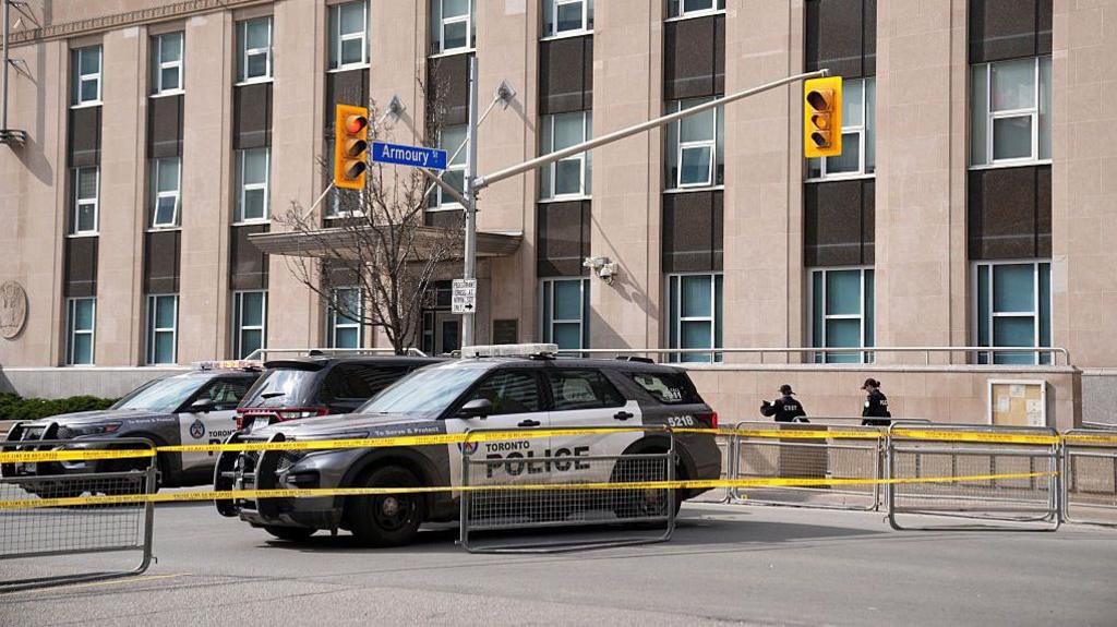 An image taken after the March shooting of the US Consulate building in Toronto. It shows three Toronto police cars behind caution tape and metal fences. Behind them is the building, which is beige and features an entrance and rows of vertical windows.