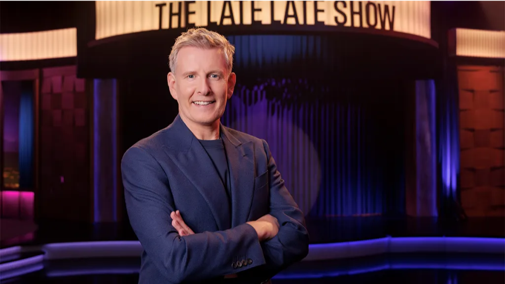 Patrick Kielty. He has blonde hair, wearing a navy suit. He is standing in front of the Late Late Show set with his arms crossed.