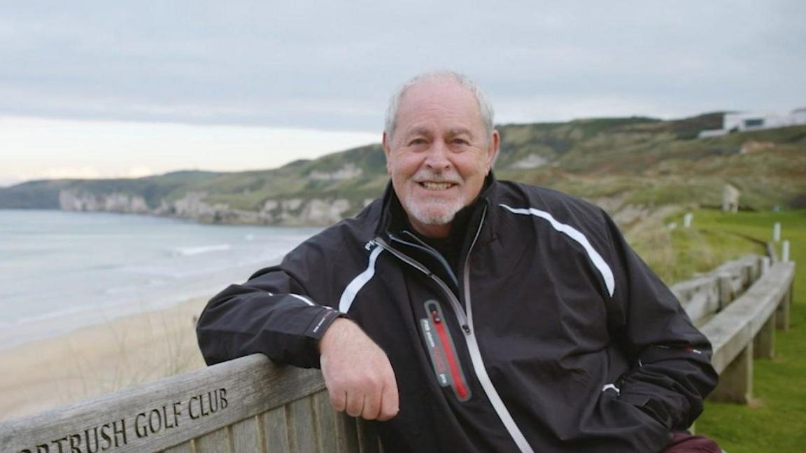 A man with short white hair smiles at the camera. He is wearing a black jacket and sitting on a bench outside. Behind him is a beach.