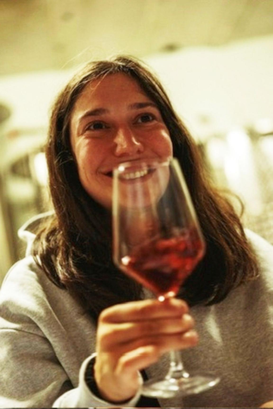 Glass containing red wine held in the foreground by a person seated indoors. The person is wearing a light-coloured sweatshirt. The glass is positioned close to the camera, with the person and a softly lit interior visible in the background.