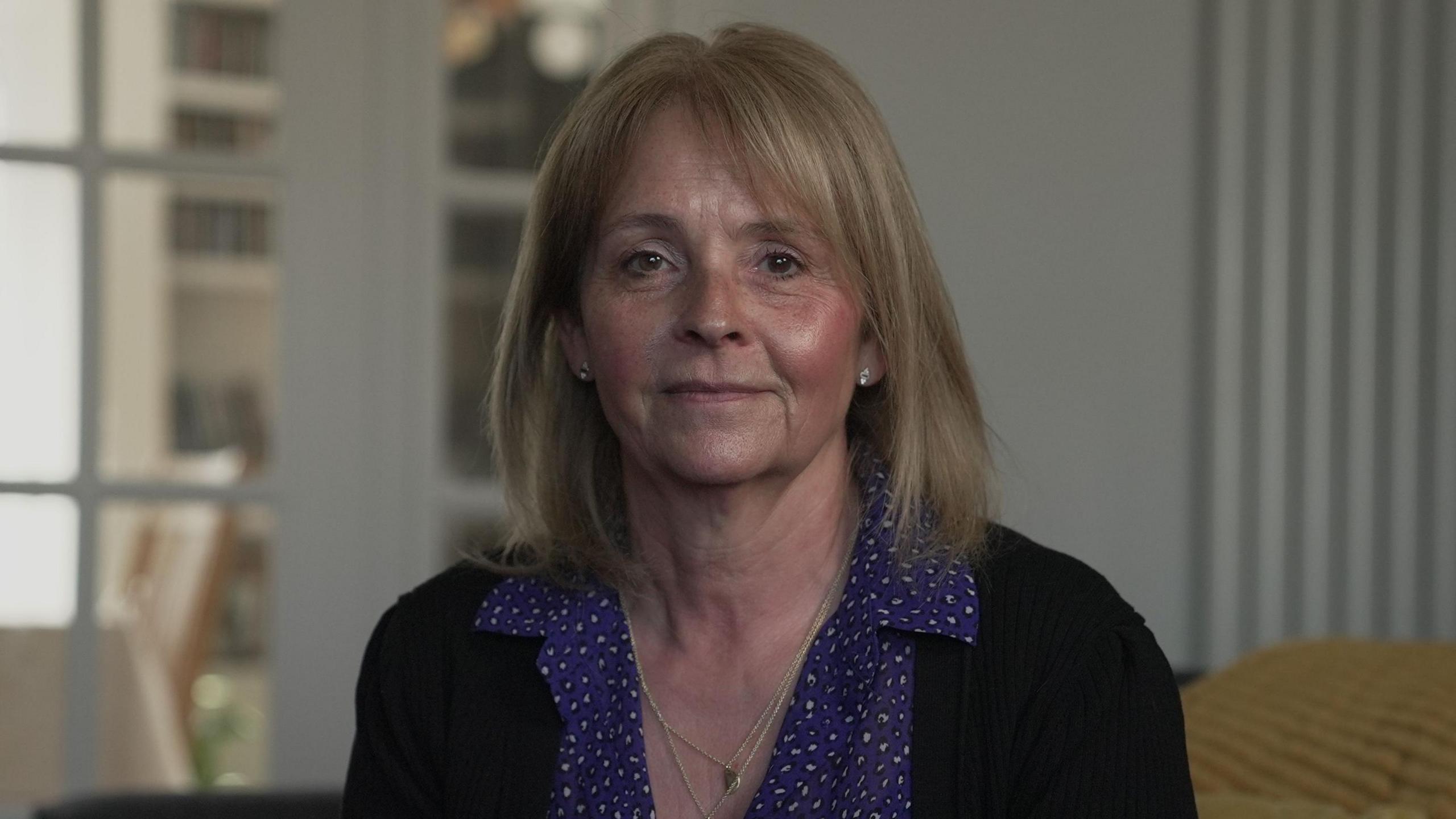Kirsten Mahoney is sat inside a house. She has shoulder length blonde hair and is smiling, looking at the camera. She is wearing two gold necklaces and stud earings in each ear. She is also wearing a pruple patterned blouse and black cardigan.