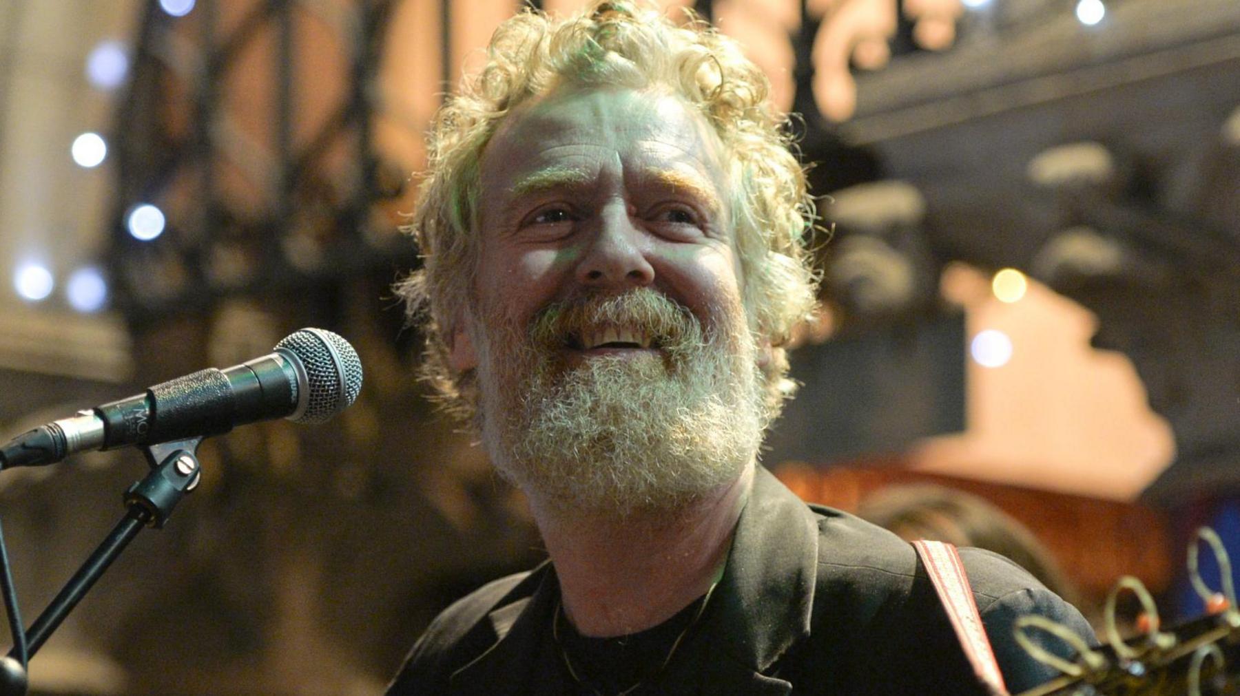 Glen Hansard is sitting in front of a microphone, he has greying red curly hair and a beard. He's smiling.