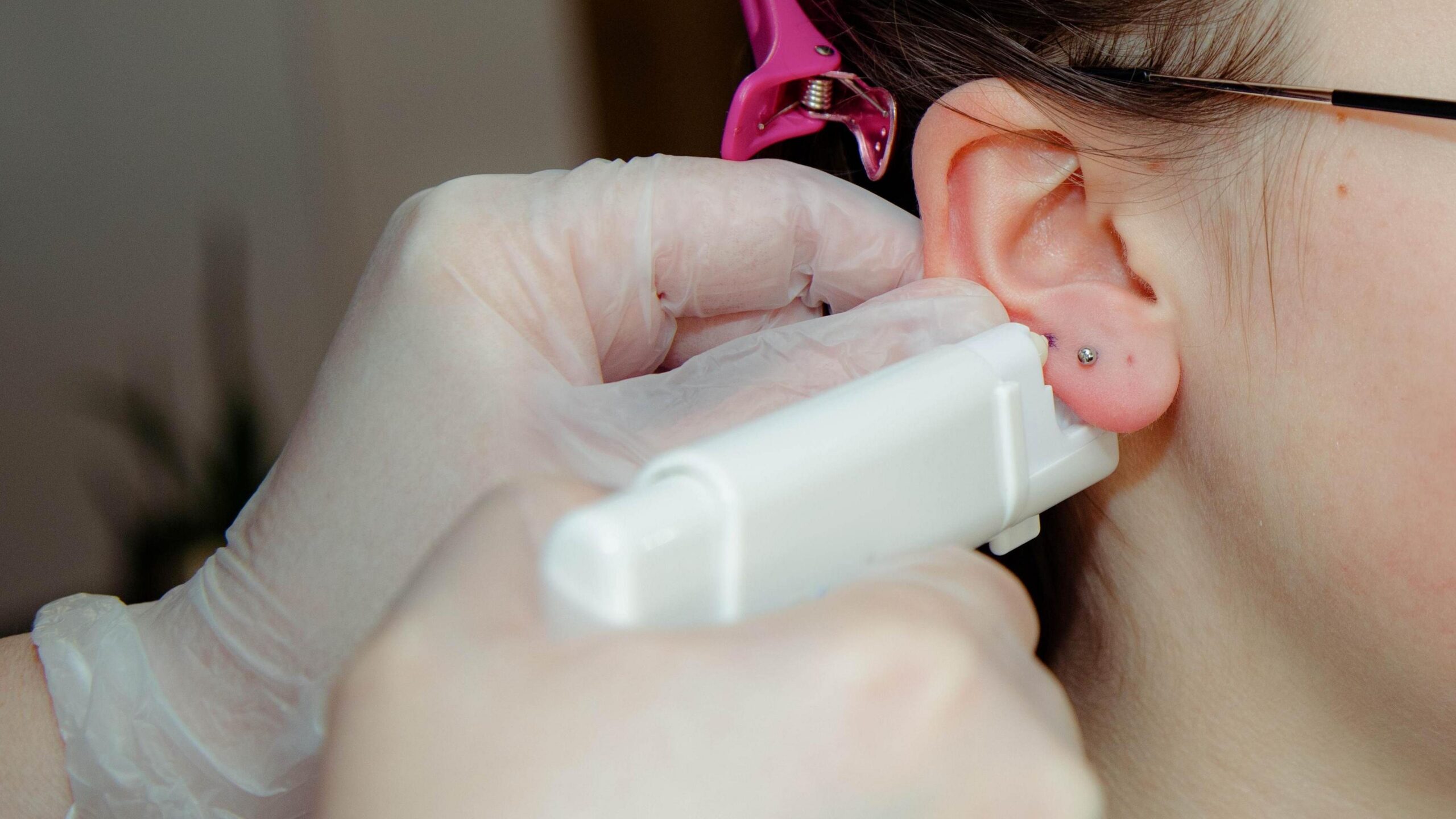 Close up of a hand of unrecognizable adult in protective gloves holding piercing gun in hand and preparing to make puncturing in earlobe of a teenage girl