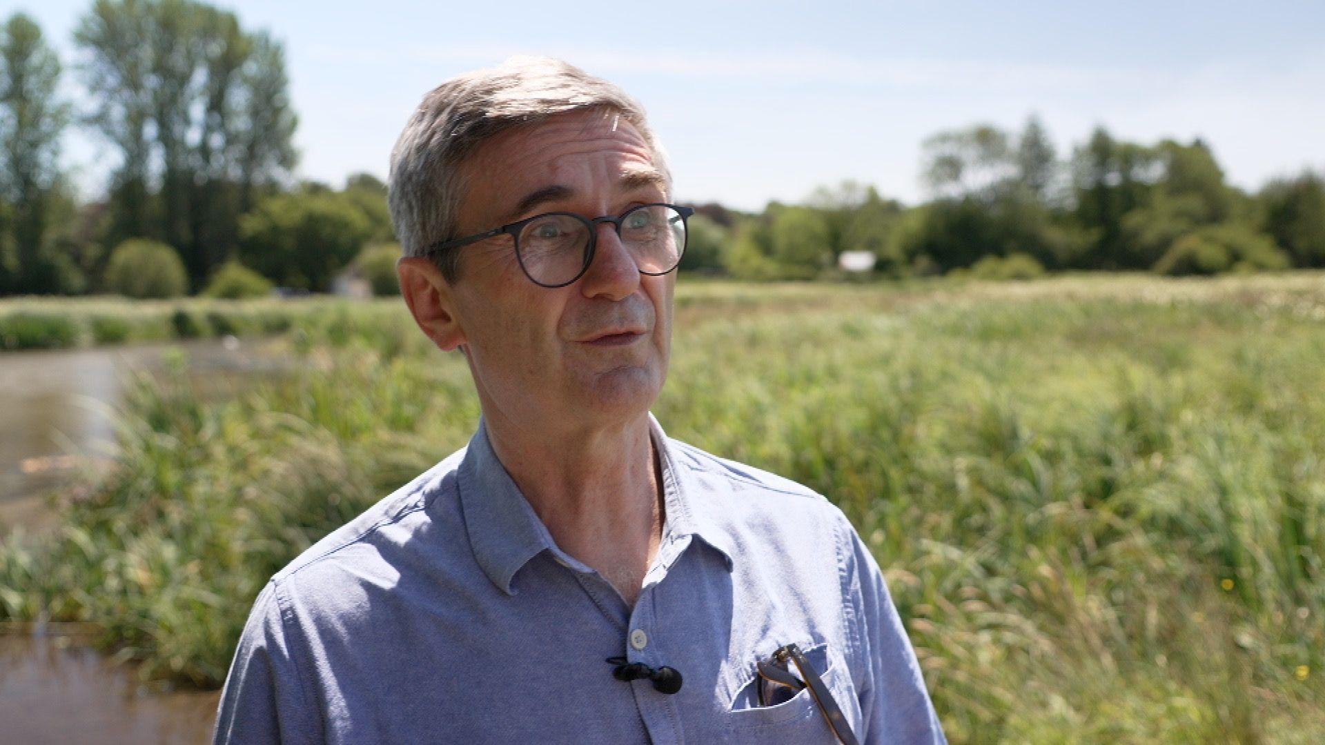 A man with grey hair, glasses and a blue shirt stands in front of water meadows with a river in the background