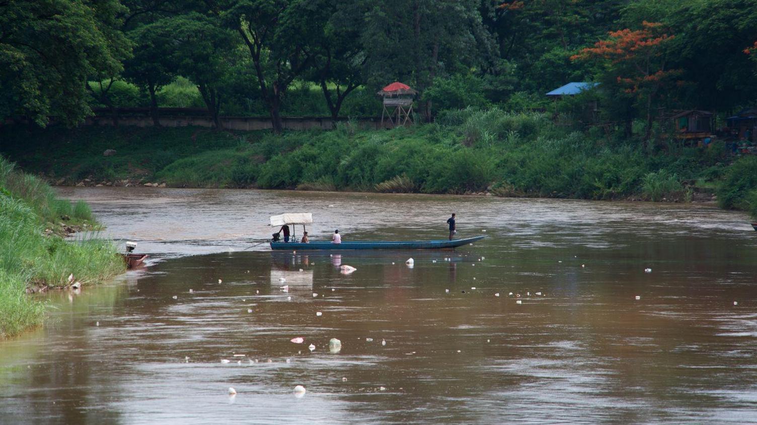 People ride a blue boat on a river with brown water, with thick foliage in the background in Mae Sot Thailand, which borders Myanmar