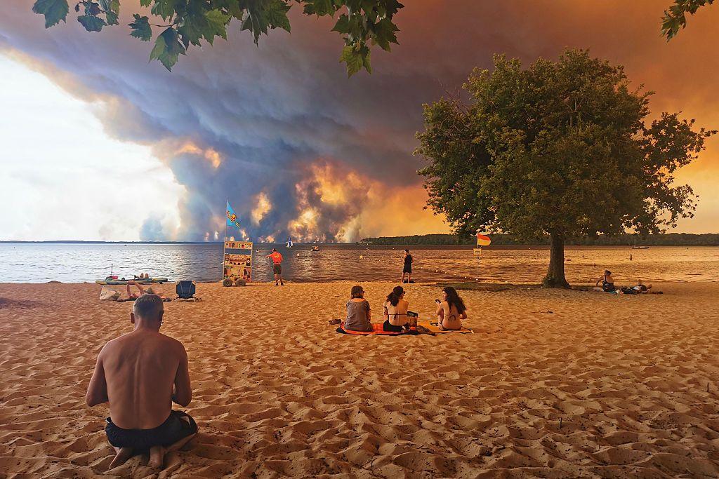 Beach goers rest on the beach of Moutchic on the shore of Lake Lacanau, southwestern France on July 24 as clouds of smoke rise into the sky from the Gironde wildfire. Huge smoke clouds and an orange sky are seen in the distance across the water, while people sit on the beach in the foreground.
