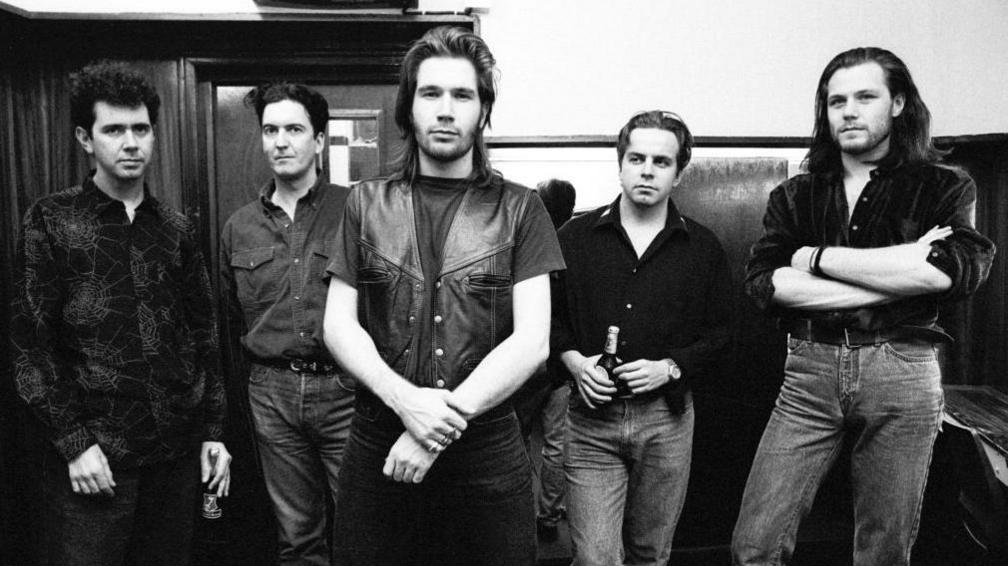Black and white photo of Del Amitri. They are standing in a hallway and are all wearing jeans and shirts. Justin Currie is also wearing a leather waistcoat