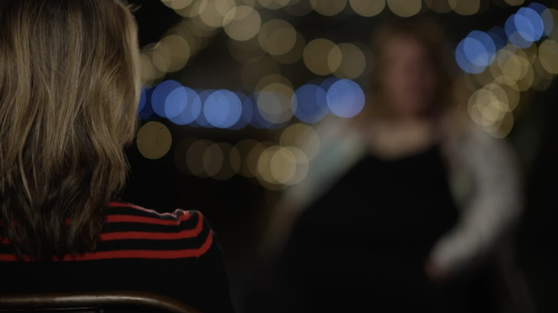 A woman with dark blonde hair and a striped red and blue top sits in a chair with her back to the camera. Another woman in a dark dress over a lighter top is sitting in a chair opposite, but her features have been blurred to protect her identity.