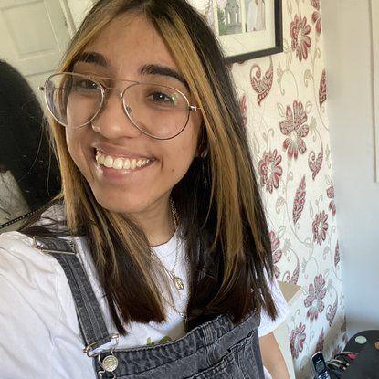 A selfie shot of Jazmin Kaur smiling on camera. She's wearing dungarees and a white t-shirt and has long brown hair and clear glasses.